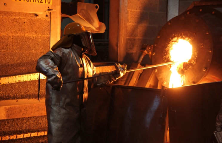 Worker in a heat-resistant suit and face shield at a smelting furnace, beside a stream of molten metal.