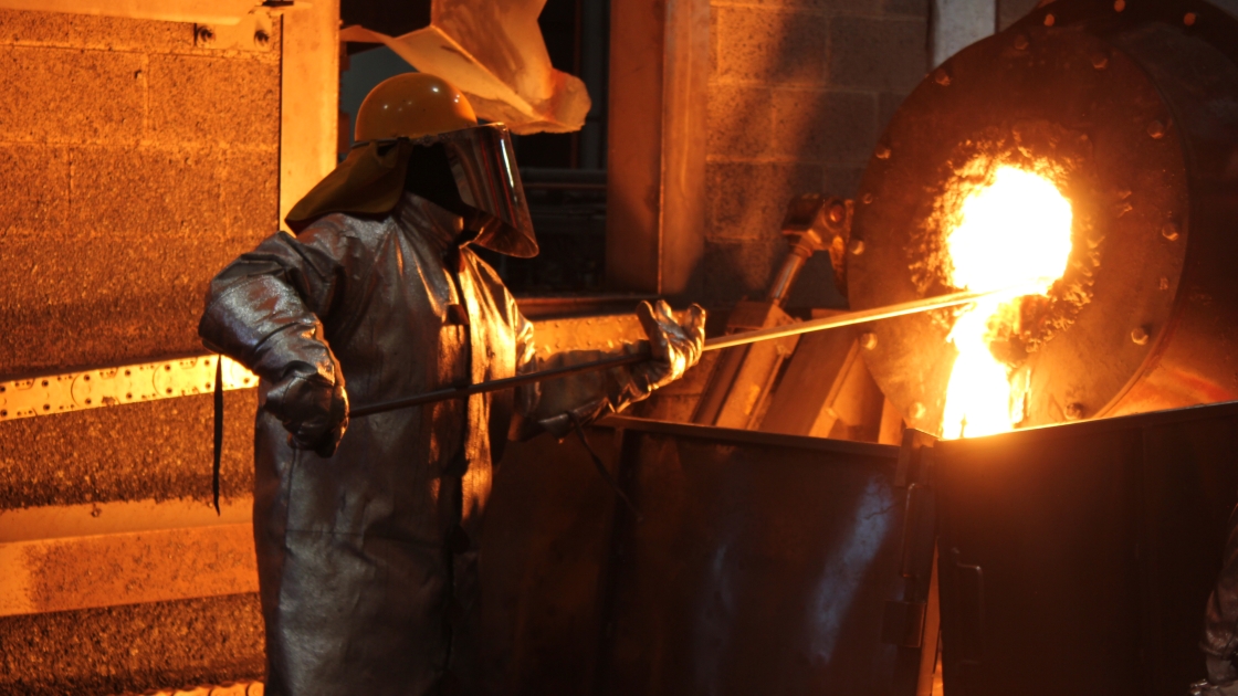 Worker in a heat-resistant suit and face shield at a smelting furnace, beside a stream of molten metal.